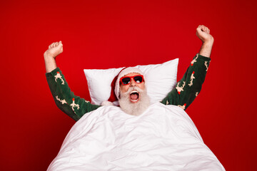 Festive Santa in cozy pajamas jumps in bed with red background celebrating Christmas and New Year...