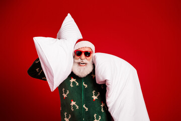 Playful santa in green reindeer robe wears red sunglasses while hugging oversized pillows against a...