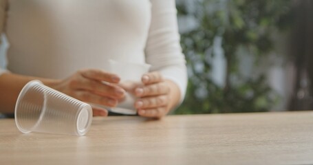 A woman holds a transparent plastic cup, with another cup lying on a wooden table in a blurred background.