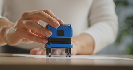 A close up image of a woman hand using a blue stamp on a sheet of paper, focusing on office tools.