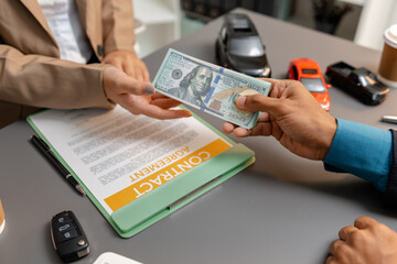 A businessperson and client are reviewing and signing a car sales contract at a desk, with small model cars, a coffee cup, and documents around them, symbolizing vehicle purchase negotiation.