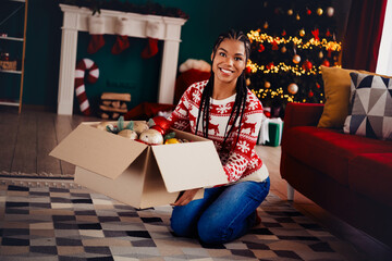 Fun festive moment a woman kneels with a box of ornaments at home preparing for Christmas tree...