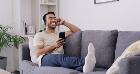 Obraz premium Relax, phone and headphones with man on sofa for streaming, radio station and audio playlist. Podcast subscription, music and media platform with person in living room of home for connection