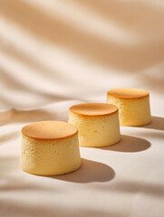 Three soft airy sponge cheesecakes with golden browned tops placed on textured fabric, bathed in warm natural light and gentle shadows, creating a minimalist elegant dessert scene.