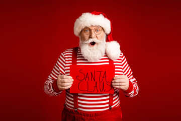 Santa Claus wearing a red hat striped shirt and suspenders holds hand written sign smiles in a festive red studio perfect for christmas advertising campaigns