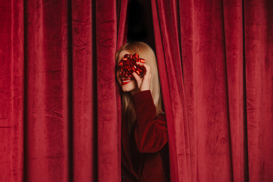 Smiling woman in stylish clothes holds a gift bow behind red velvet curtain
