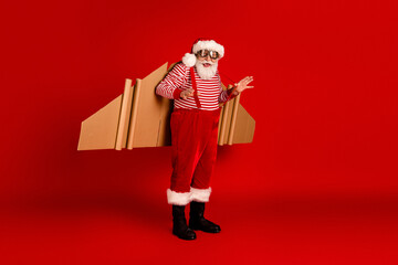 Santa in a striped red and white shirt with suspenders carries cardboard rocket wings on a vivid red studio backdrop for a festive holiday stock photo