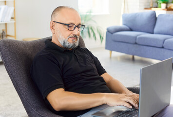 Senior man with glasses using laptop to work online or banking in living room workspace. Busy person typing on PC keyboard, working with productivity on remote freelance project from home office