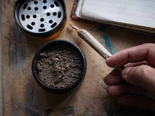 Hand holding rolled marijuana joint with grinder and cannabis herb on wooden table for recreational use