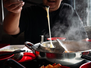 Person stretching melted cheese with chopsticks from steaming hot pot shabu divided metal pot on dining table