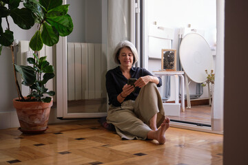 Relaxed senior woman using mobile phone at home sitting on the floor near balcony door