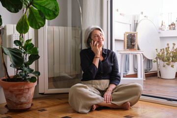 Happy mature woman using mobile phone, making call and smiling while sitting on the floor at home
