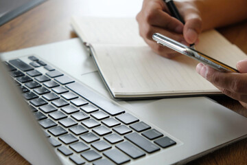 Person writing notes in notebook while holding smartphone near laptop keyboard, multitasking with focus and planning for business or study purpose