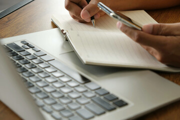 person holding smartphone while writing notes in notebook beside laptop, multitasking with focus and planning for business or study purposes