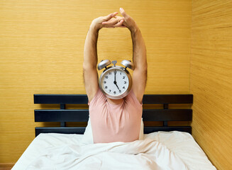 Happy man with alarm clock instead of head stretching on bed at home