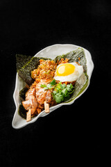 A close-up of an Asian-inspired dish served in a white bowl. The meal features fried rice, skewered chicken, a sunny-side-up egg with sesame seeds, cilantro, and nori seaweed.