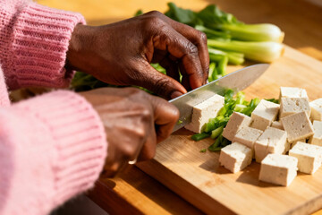 Close-up of mature African woman’s hands cutting tofu and green vegetables, soft morning light, pink knit sleeves, concept of hormonal balance and plant-based diet.
