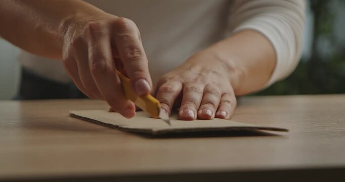 Hands woman cutting a piece of cardboard with a utility knife on a wooden table. Careful craftsmanship in focus.