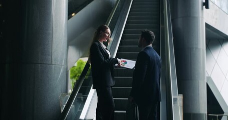 Formal man and woman executives stand discussing colorful graph and data analysis on paper documents while moving up on escalator of modern corporate building - Powered by Adobe