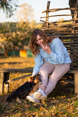 A woman sits on a rustic wooden bench, surrounded by the warm glow of golden autumn. She smiles while petting her dog, enjoying the peaceful beauty and cozy charm of the fall season.