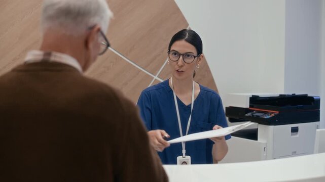 Medium shot of female medical receptionist asking aged Caucasian man to sign document in modern clinic