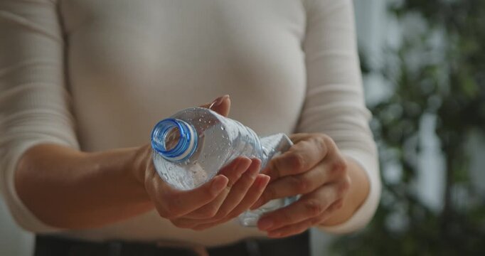 A woman hands twisting empty plastic water bottles on a wooden table. A bright room with simple decor and a green plant in the background.