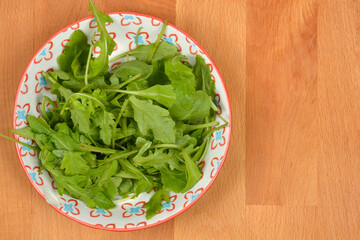 A small, decorative bowl full of fresh green arugula leaves is positioned on the left side of a light wooden background in an overhead shot