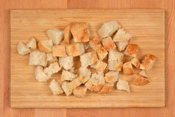 Small, roughly diced cubes of bread are scattered on the light wooden cutting board, set on a wooden background