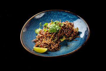 A dish of dark noodles with sesame seeds, fresh herbs, and a lime wedge, served in a blue bowl against a black background.