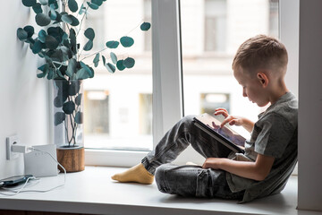 A 6-year-old boy is sitting on a windowsill with a tablet. A child is playing with a phone. The dangers of gadgets for children. Protect your child from smartphones. Children and gadgets.
