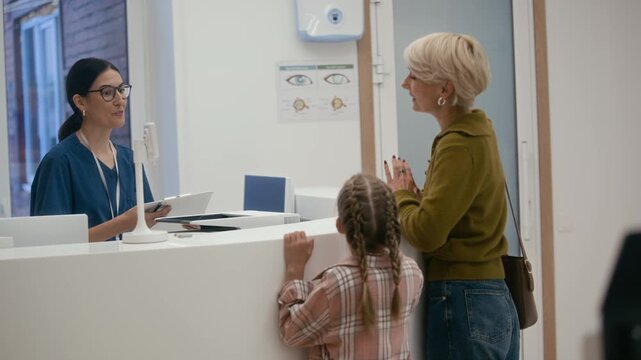 Medium long shot of female medical administrator in uniform asking to sign form while consulting middle aged woman and her daughter at clinic reception