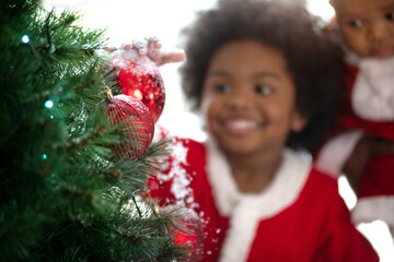 Happy African child boy Santa costume decorating red ball on Christmas tree at home, He noticed...
