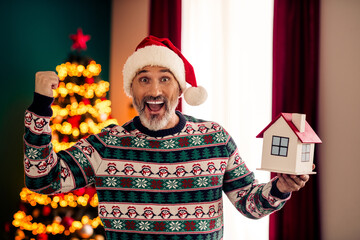 Happy senior man in festive sweater holding small house model embracing holiday cheer with...