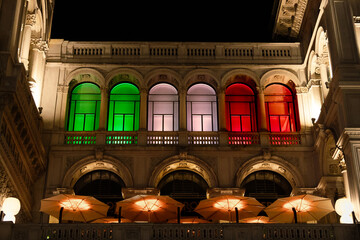Night view of Galleria Vittorio Emanuele II with windows illuminated in the colors of the Italian flag, green, white, and red.