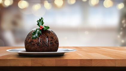 A low angle view of a traditional Christmas pudding on a round plate with a sprig of holly, berries on a wooden table against a light festive background of Christmas tree lights.