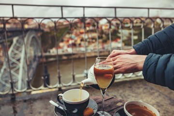 A person pours tea from a white teapot into a black teacup and a tall glass. The Dom Luis I bridge and buildings of Porto are visible in the background