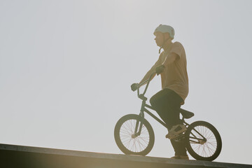 Fototapeta premium Teenager riding BMX bike on ramp outdoors wearing helmet, looking forward with focused expression, sunlight creating strong backlight, engaging in extreme sport activity