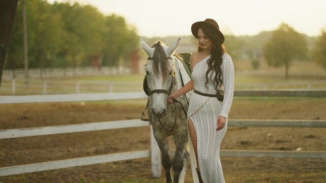 Woman in hat holding reins next to gray horse on ranch. Woman with horse concept.