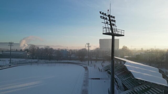 Stadion sportowy zimą widziany z drona. Pokryte śniegiem trybuny i boisko tworzą malowniczy, spokojny krajobraz. Ujęcie z lotu ptaka ukazuje symetrię obiektu i kontrast między bielą śniegu a otoczenie
