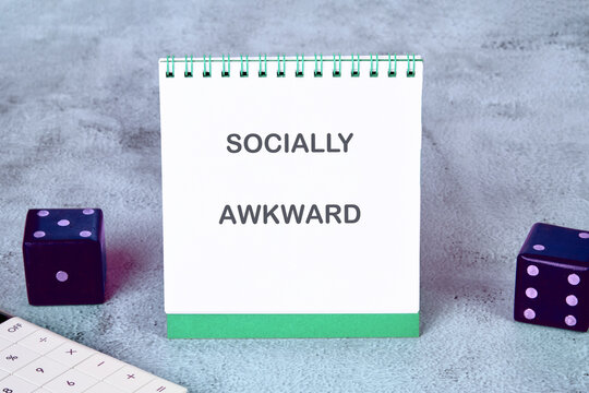 A notepad with the words socially awkward stands prominently on a soft gray surface beside two colorful dice. This scene invites reflections on personal experiences and humorous situations