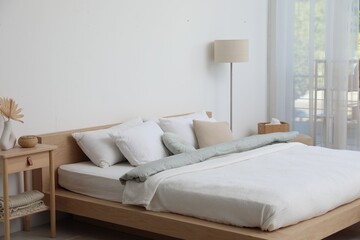 Cozy large bed with linens, wooden nightstand and lamp near white wall in bedroom. Interior design