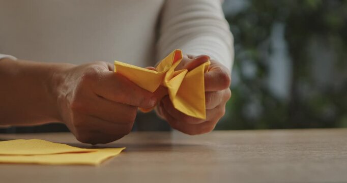 A woman hands crumple a yellow paper napkin on a wooden table surface. The room has a light atmosphere with a simple interior and a green plant in the background.