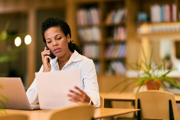 African-American Woman Working in Library Office, On Phone and Reviewing Documents for Work Busy...