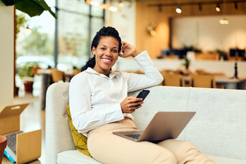 African-American Woman Relaxing in Modern Office Lobby with Laptop and Phone, Smiling