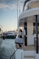 Elegant yacht docked in a marina with teak deck and polished railings. A woman stands on the deck, overlooking the calm water and nearby boats under a clear sky at Limassol Marina.