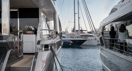 Two women standing on the deck of a luxury yacht docked in a marina. The calm blue water reflects the boats, with sailboats and yachts visible in the background under a clear sky at Limassol Marina.