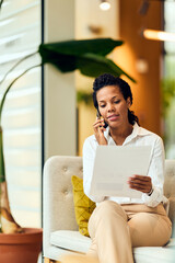 African-American Woman On Phone Reviewing Document In Modern Office Lounge Setting During Casual...