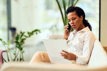 African-American Woman On Phone While Reviewing Document In Modern Office Setting During Workday