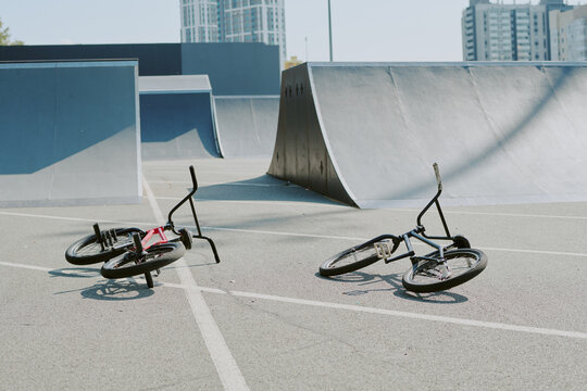 Two BMX bikes lying on ground at outdoor skatepark with modern ramps and urban buildings in background, suggesting recent activity by teenagers or young adults - Powered by Adobe