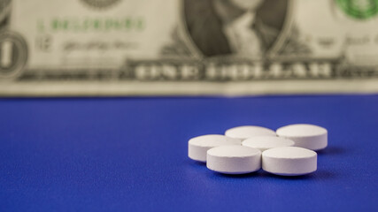 White pills on blue surface near a dollar bill represent health and finance concepts
